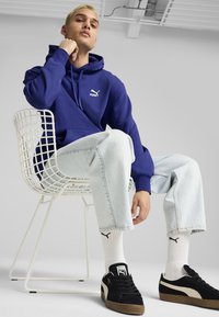 Felpa blu con logo Puma, abbinata a pantaloni in denim blu chiaro. Sneakers nere con dettagli bianchi, seduto su una sedia di filo bianco.