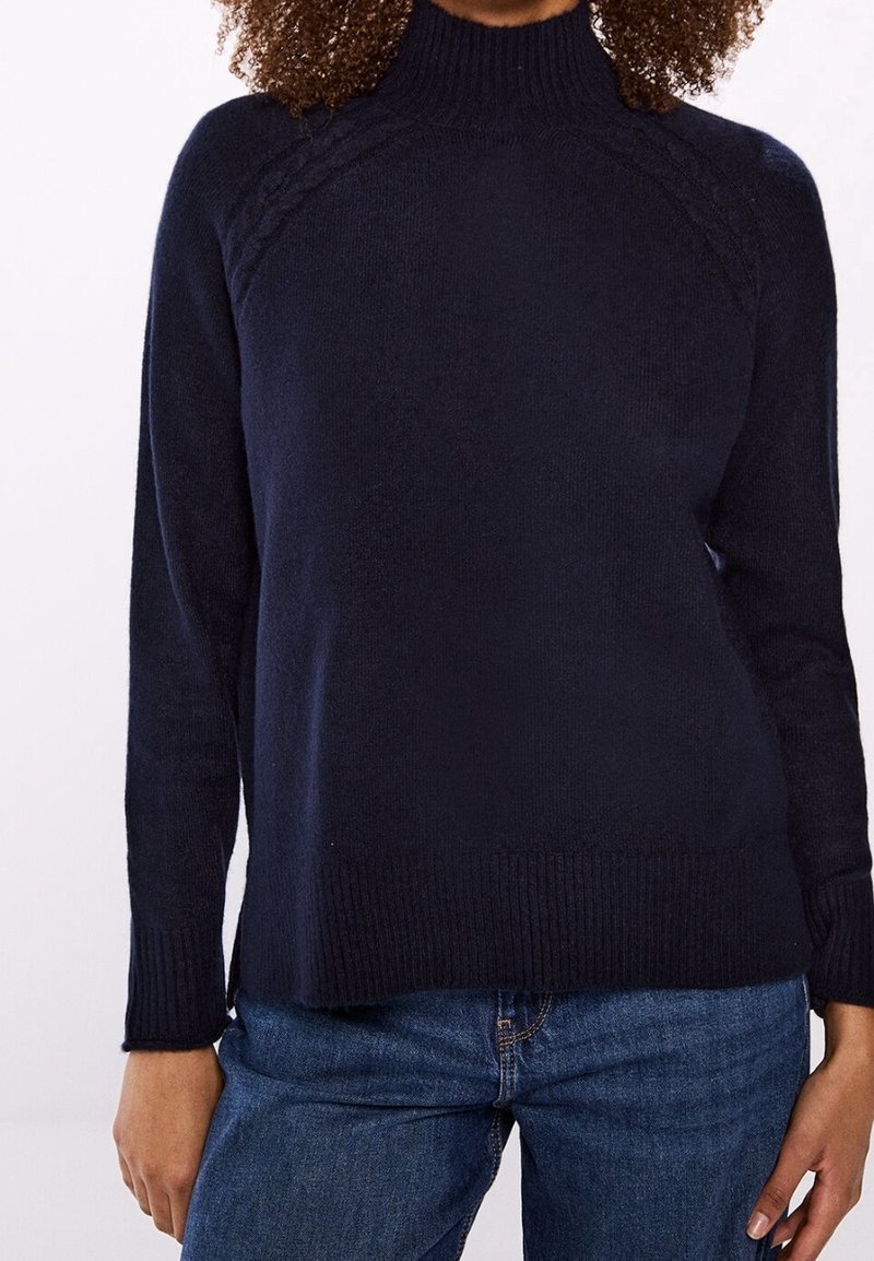 Femme aux cheveux bouclés portant un pull en tricot bleu marine à col montant et un jean bleu, debout devant un fond uni.