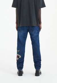 Jeans de mezclilla en azul con un corte relajado, que presentan un parche decorativo en la pierna izquierda con elementos coloridos y un detalle de costura en contraste.