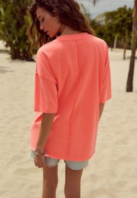 Maglietta oversize in cotone rosa neon con maniche corte, caratterizzata da una cucitura centrale e una vestibilità comoda, abbinata a pantaloncini di denim chiaro.