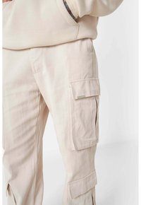 Beige Cargo-Hosen aus weichem Stoff; verfügen über seitliche Taschen, einen lockeren Schnitt und sichtbare Nähte. Akzentuiert durch einen subtilen Glanz.