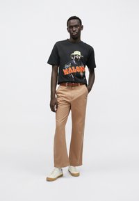 Black t-shirt with graphic print, tan wide-leg trousers, brown belt, and white sneakers; presented against a neutral background.