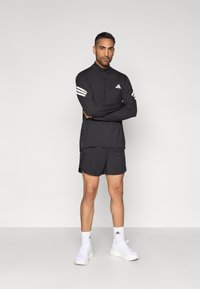 Sort atletisk langærmet trøje med lynlåshals og tre hvide striber på ærmerne, kombineret med sorte shorts og hvide sneakers.