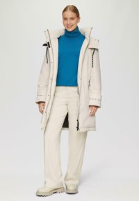 Beige puffy jas met een hoge kraag, rits- en knoopsluiting, gecombineerd met een blauwe coltrui en crèmekleurige broek. Bevat zwarte koordaccenten.