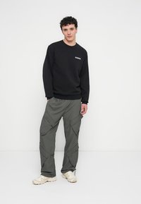 Felpa nera con logo sul petto, abbinata a pantaloni cargo grigi a taglio largo con multiple tasche e sneakers beige.