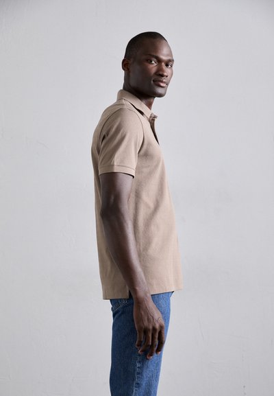 Polo beige avec un tissu texturé, à manches courtes et avec un col classique. Porté avec un jean en denim bleu, vu de côté.