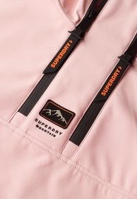Roze buitenjack met zwarte ritsen en trekkoorden. Bevat een "SUPERDRY" logo en een grafische patch van een berg op de borst.