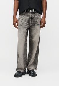 Άνδρας που φοράει φαρδιά ξεθωριασμένα μαύρα-γκρι τζιν, μαύρο πουκάμισο μέσα, μαύρη ζώνη με ασημένια αγκράφα και μαύρα αθλητικά παπούτσια σε λευκό φόντο.