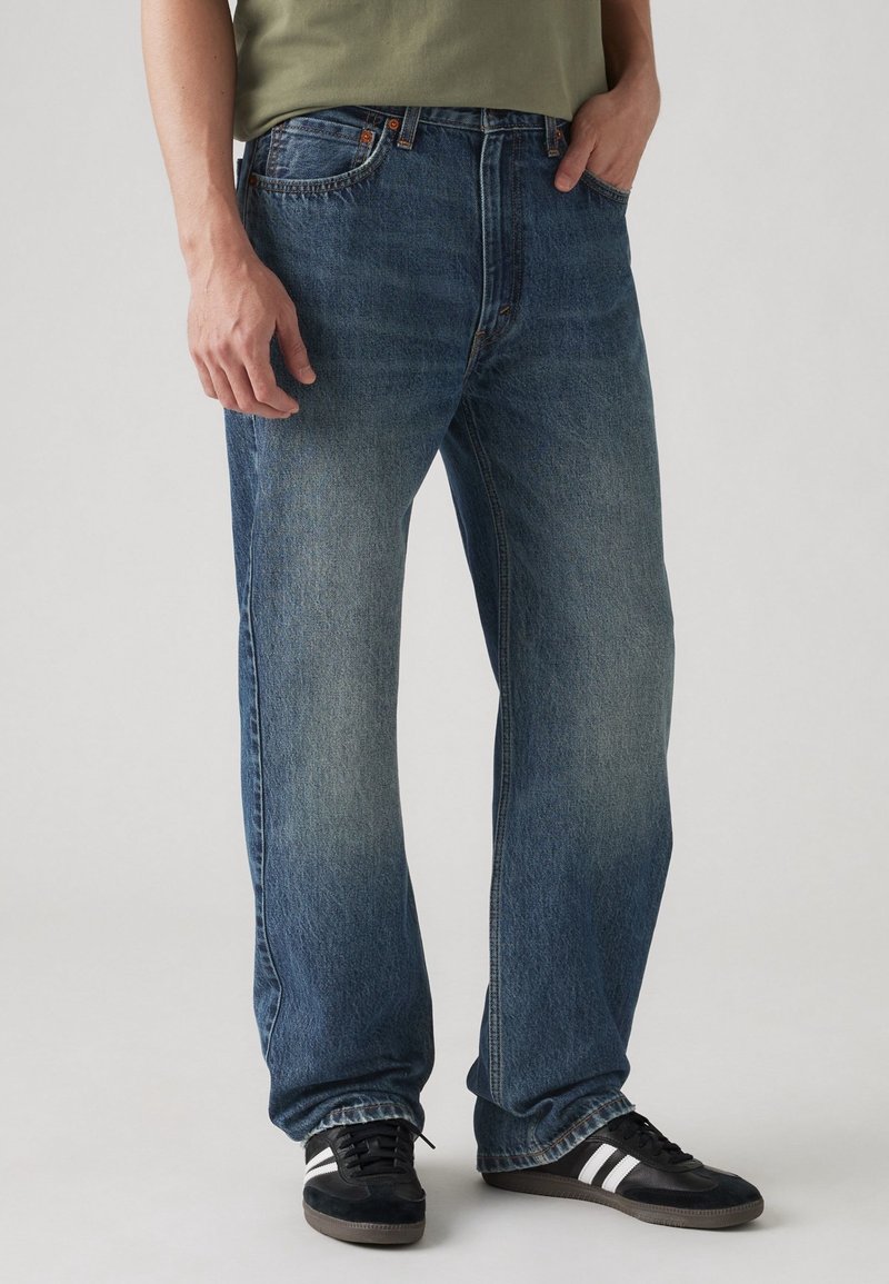 Homme portant un jean bleu délavé droit et des baskets noires à rayures blanches, debout avec une main dans la poche.