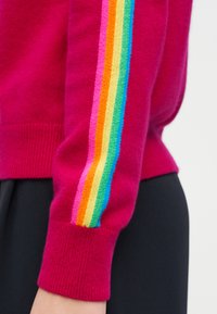 Μανίκι πουλόβερ σε ματζέντα με μανσέτα και κάθετες ρίγες ουράνιου τόξου σε ροζ, πορτοκαλί, κίτρινο, πράσινο και μπλε, φορεμένο πάνω από μαύρο παντελόνι.