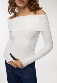 Biała prążkowana bluzka bez rękawów z długimi rękawami, z wyprofilowanym dekoltem i dopasowanym krojem, zestawiona z ciemnoniebieskimi dżinsami.