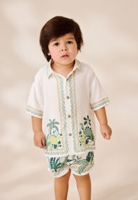 Baltas trumpomis rankovėmis marškinėlis su žaliomis botaninėmis spaudomis, dekoratyviniais mygtukais ir raštuotu apvadu; derinamas su atitinkančiomis šortais su panašiais raštais.