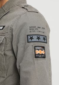 Olivengrøn militærstil jakke med knappet krave, patchdetaljer på ærmerne og fremtrædende brandlogo på frontlommen.