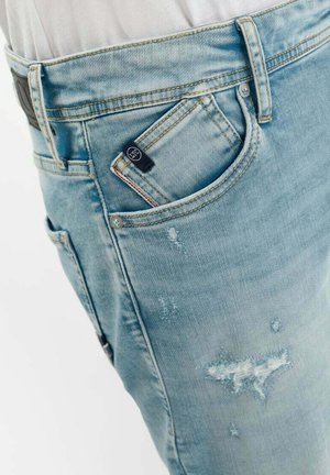 Jean en denim délavé bleu clair avec un portefeuille partiellement visible dans la poche avant droite sur fond blanc.