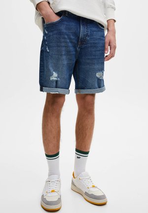 Personne portant un short en denim bleu retroussé avec des détails usés, des chaussettes blanches à rayures vertes et des baskets blanches avec des accents jaunes.