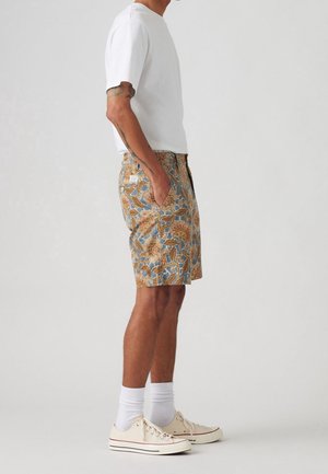 Mann in einem weißen T-Shirt, mit blumengemusterten Shorts mit Taschen, weißen Socken und beigen Leinenschuhen, der seitlich vor einem neutralen Hintergrund steht.