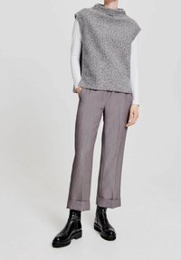 Person klädd i en grå texturerad ärmlös stickad väst över en vit långärmad topp, grå uppvikta byxor och svarta kraftiga stövlar, stående med händerna i fickorna.