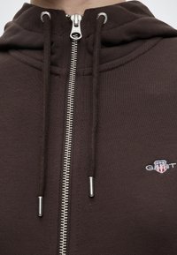 Gros plan sur un sweat à capuche marron à fermeture éclair métallique, cordons avec embouts métalliques et un petit logo GANT brodé sur la poitrine.
