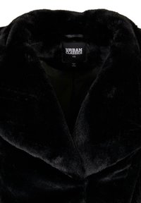 Melns mākslīgā kažoks ar plašu apkakli. Tam ir mīksta tekstūra un minimālisma interjers. Redzama birka: "URBAN CLASSICS XS".