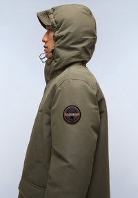 Giacca isolante khaki con cappuccio, dotata di una toppa rotonda sul maniche sinistro. Texture liscia, design robusto e coulisse regolabile.