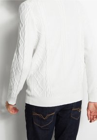 Pull en tricot blanc avec des motifs de câbles texturés et des poignets côtelés. Assorti à un jean bleu foncé avec des détails de couture jaune.