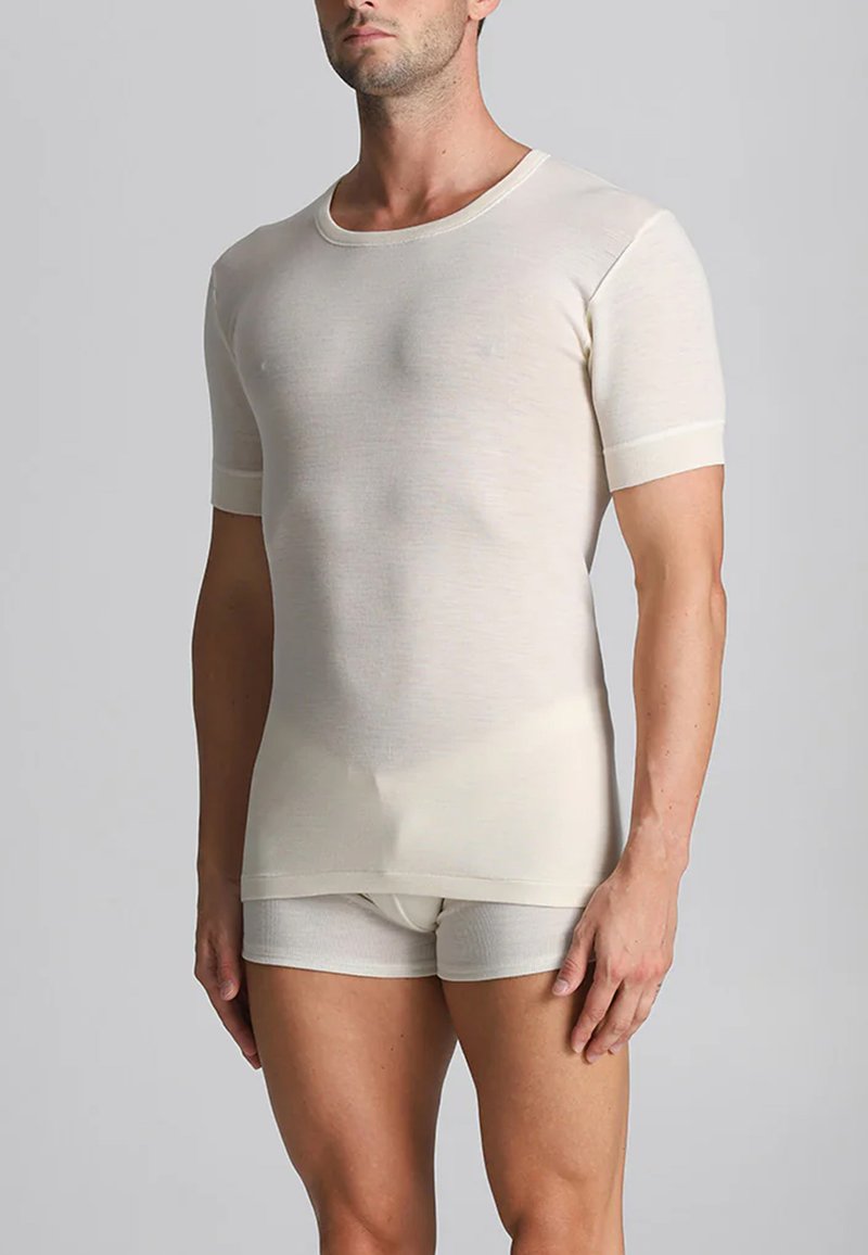 Uomo che indossa una camicia a maniche corte aderente color crema e slip coordinati che si erge contro uno sfondo grigio uniforme.
