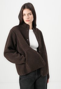 Brun fleecejakke med myk tekstur, trykknap-lukking og åpen krage-design. Har sidelommer og en avslappet passform.