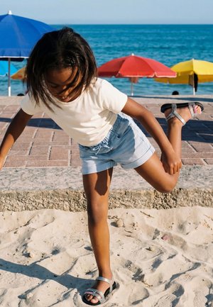 Bambino con camicia bianca e pantaloncini blu che si equilibra su una gamba sulla spiaggia di sabbia vicino a ombrelloni colorati e all'oceano.