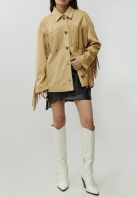 Beige jas met franjes en knoopsluiting, met een kraag, gecombineerd met een zwarte rok met franjes en kniehoge witte laarzen.