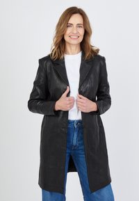 Schwarzes Lederjacke mit Spitzkragen, vorderen Knöpfen und zwei Taschen. Kombiniert mit einem weißen Hemd und blauen Jeans. Glatte Textur.