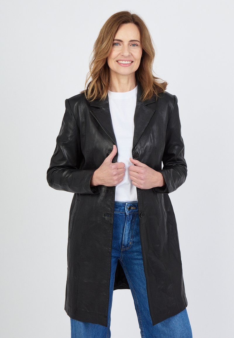 Schwarzes Lederjacke mit Spitzkragen, vorderen Knöpfen und zwei Taschen. Kombiniert mit einem weißen Hemd und blauen Jeans. Glatte Textur.