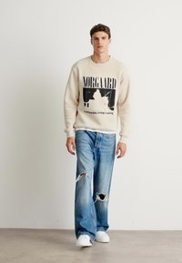 Beige sweatshirt with black graphic text and image on front, paired with light blue ripped jeans and white sneakers. Smooth texture.