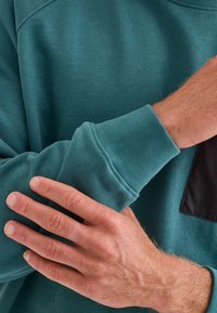 Sudadera turquesa con puños acanalados, textura suave y un acento en forma de bolsillo negro. Mano descansando sobre la manga que muestra una detallada textura de la piel.