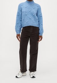 Suéter de punto azul claro con cuello alto, que presenta un patrón texturizado, combinado con pantalones de pana marrón oscuro y zapatillas blancas.