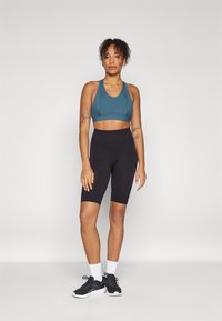 Teal sports-BH og svarte høytaljede sykkelshorts. BH-en har racerskærf design. Modellen har på seg svarte treningssko og hvite ankelstrømper.