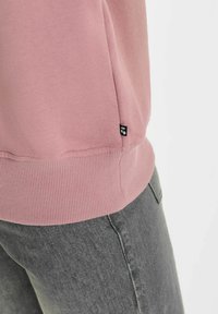 Lichtroze sweatshirt met geribbelde zoom en manchetten, met een klein zwart logotag aan de zijkant. Gemaakt in combinatie met grijze spijkerbroek.