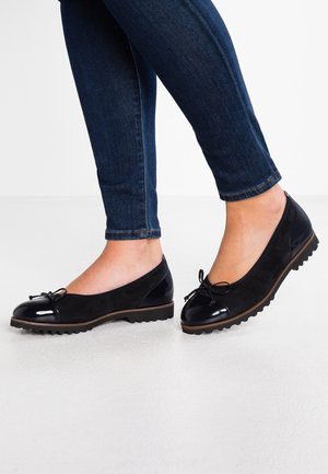 Ben i mørkeblå jeans iført sorte balletsko med skinnende tåhætte, små sløjfer og teksturerede såler på en hvid overflade.