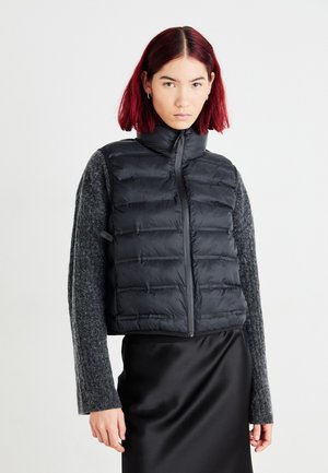 Zwarte puffer vest met hoge kraag, gewatteerd ontwerp en ritssluiting aan de voorkant. Gecombineerd met een grijs langslavend shirt en een zwarte satijnen rok.