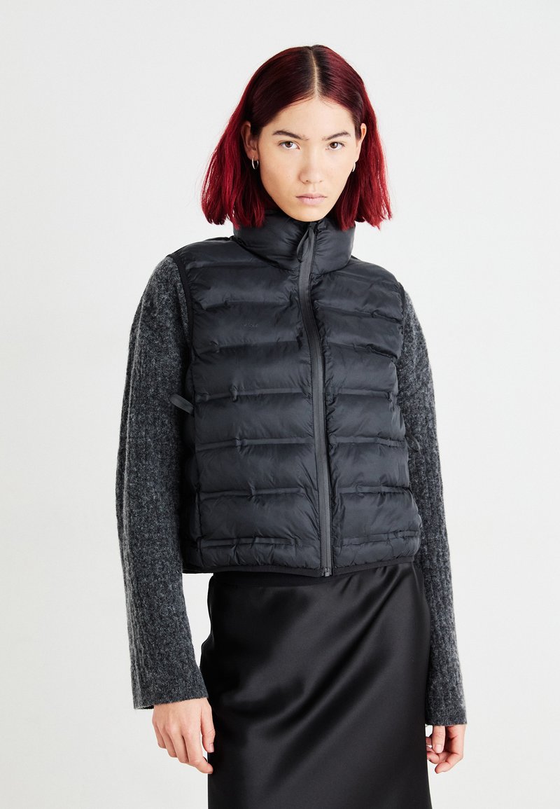 Zwarte puffer vest met hoge kraag, gewatteerd ontwerp en ritssluiting aan de voorkant. Gecombineerd met een grijs langslavend shirt en een zwarte satijnen rok.