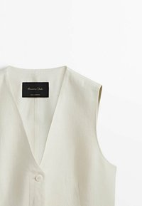 Gilet sans manches de couleur crème avec un profond col en V, une fermeture par un bouton blanc unique et une étiquette de marque noire à l'intérieur du col.
