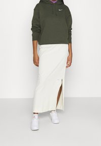 Oliwkowa bluza z kapturem z logo na przodzie, biała maxi spódnica z bocznym rozcięciem i białe sneakersy. Prosty, casualowy strój z tekstylnymi detalami.