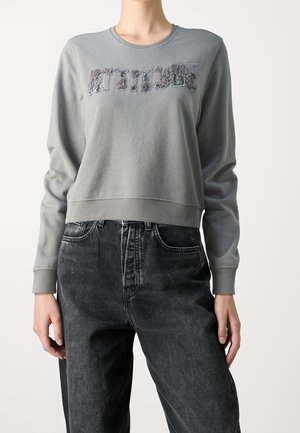 Szara krótka bluza z długim rękawem z fakturowanym napisem „ATTITUDE” w zestawieniu z wysokim stanem wyblakłych czarnych dżinsów.