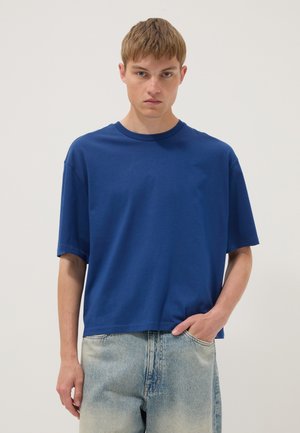 Joven con una camiseta azul lisa de manga corta y vaqueros azul claro descoloridos, de pie con una mano en el bolsillo frente a un fondo liso.