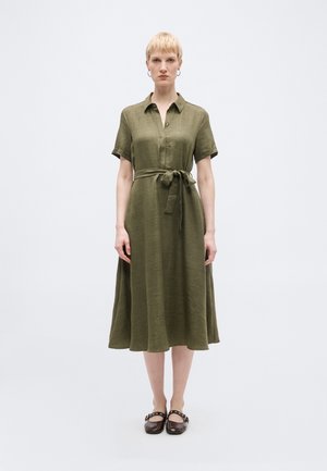 Γυναίκα με κοντά ξανθά μαλλιά που φοράει μεσοφόρι ελιάς με κοντά μανίκια, γιακά, ζώνη στη μέση και σκούρα καστανά παπούτσια Mary Jane.