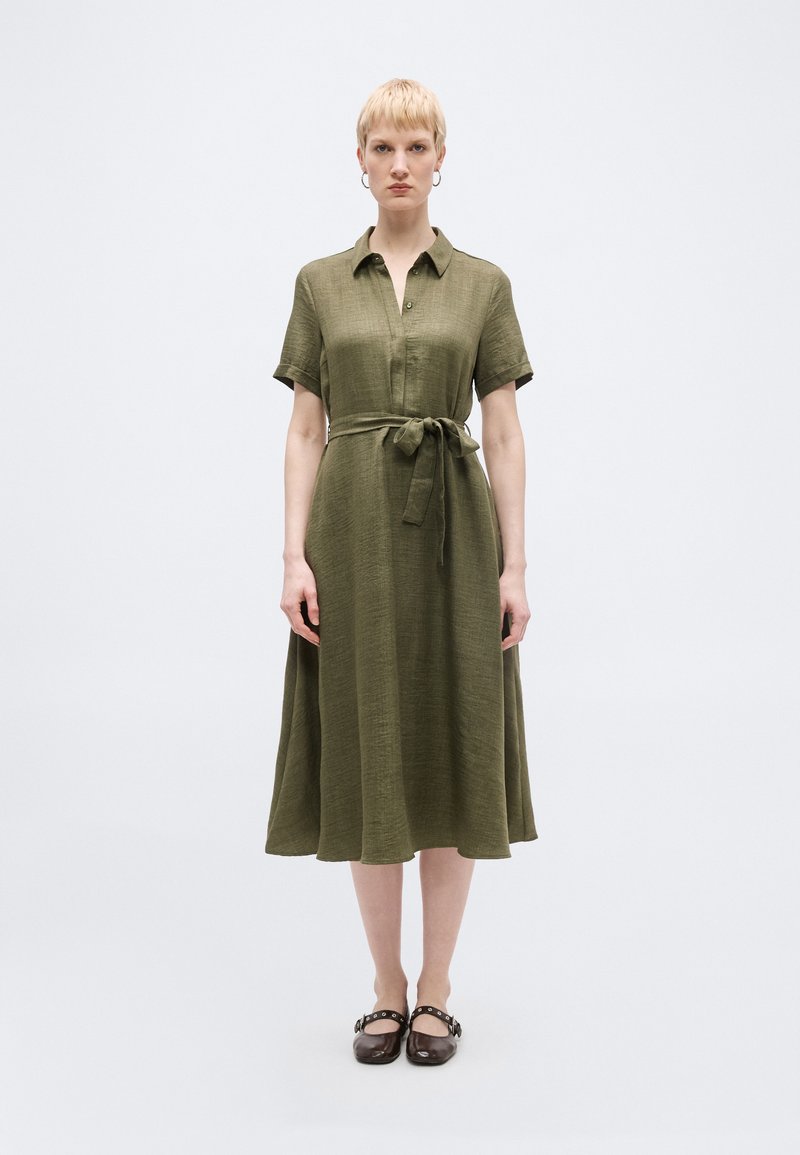 Γυναίκα με κοντά ξανθά μαλλιά που φοράει μεσοφόρι ελιάς με κοντά μανίκια, γιακά, ζώνη στη μέση και σκούρα καστανά παπούτσια Mary Jane.