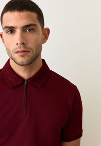 Bordo polo marškinėliai su tekstūruotu, raštuotu užtrauktuko apykakliu ir trumpomis rankovėmis, pagaminti iš sklandžios, lengvos medžiagos.