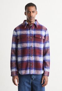 Red and blue plaid flannel shirt with a button-up front, two chest pockets, and a pointed collar. Features a textured fabric.
