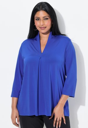Blauwe, lange mouwen top met een V-hals en plooidetails, gemaakt van een gladde, gestructureerde stof. Gekleed met zwarte broek, tegen een witte achtergrond.