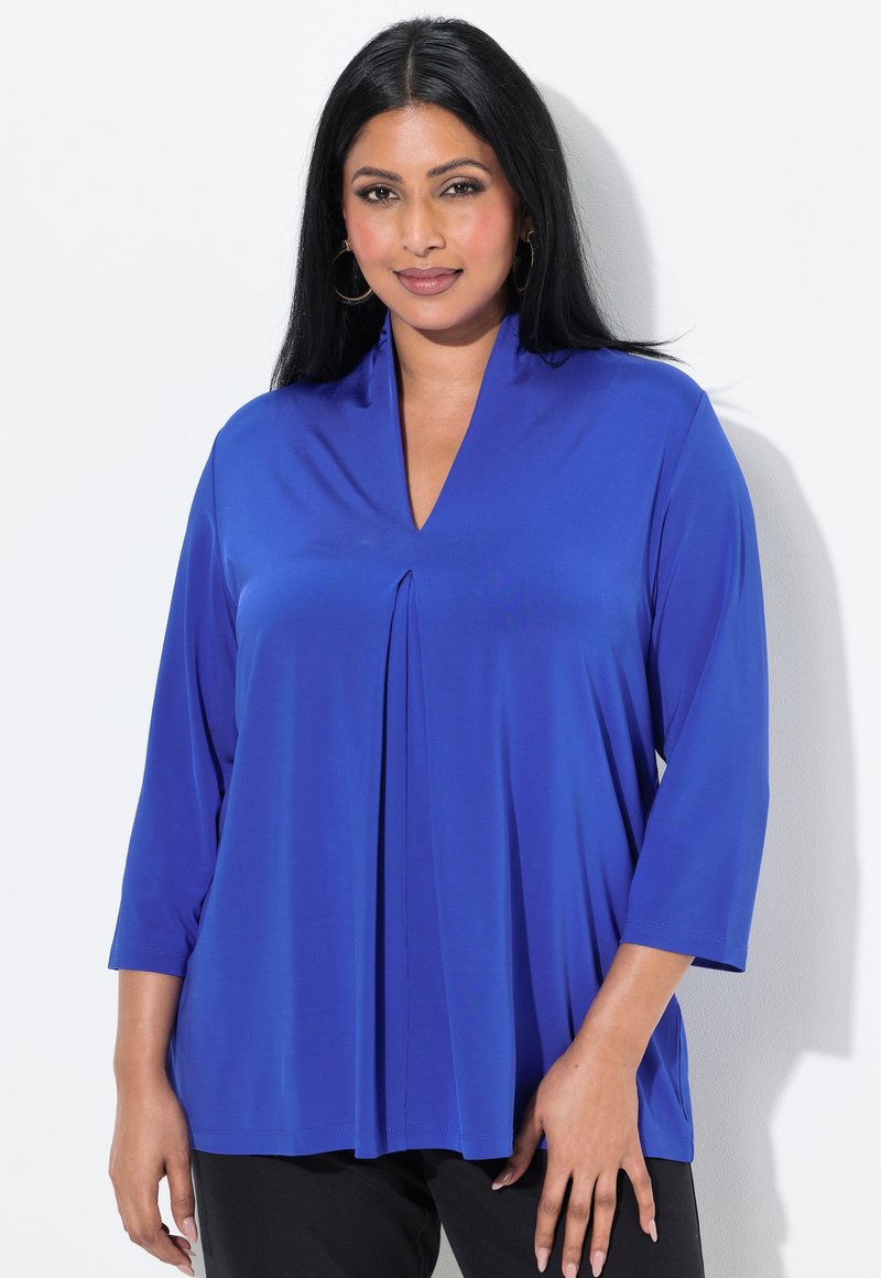 Top blu, a maniche lunghe, con scollo a V e dettagli plissé, realizzato in un tessuto liscio e testurizzato. Abbinato a pantaloni neri, su uno sfondo bianco.