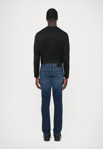 Camisa negra de manga larga y vaqueros de mezclilla azul oscuro vistos desde atrás. Los vaqueros presentan un corte clásico, con bolsillos traseros y detalles mínimos.